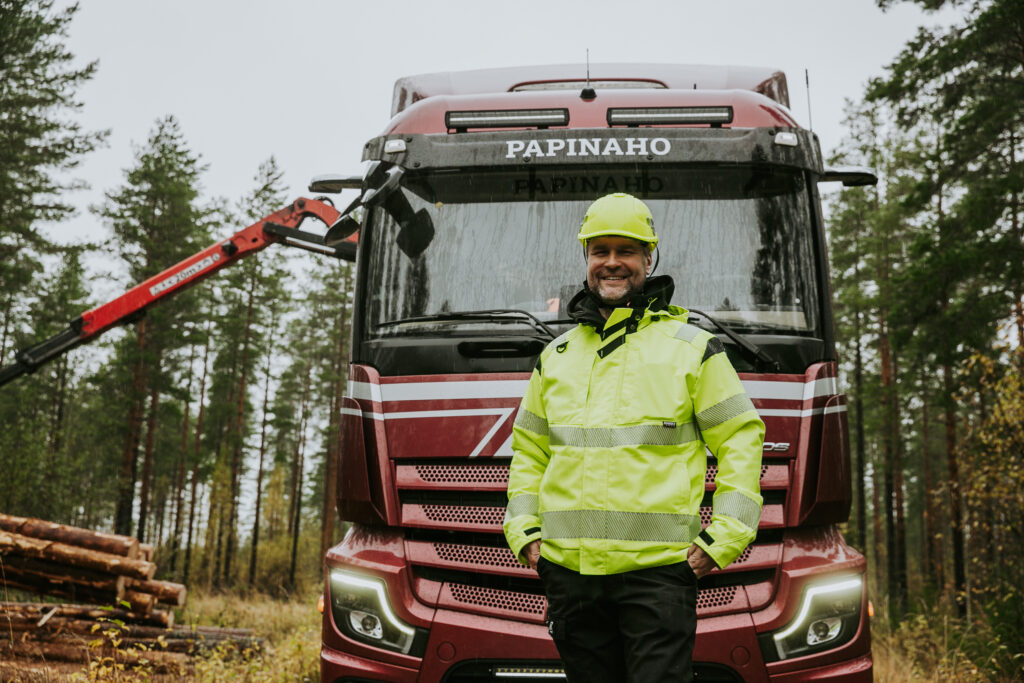 Mies huomiotakissa ja turvakypärä päässä seisoo metsässä. Taustalla puutavara-auto